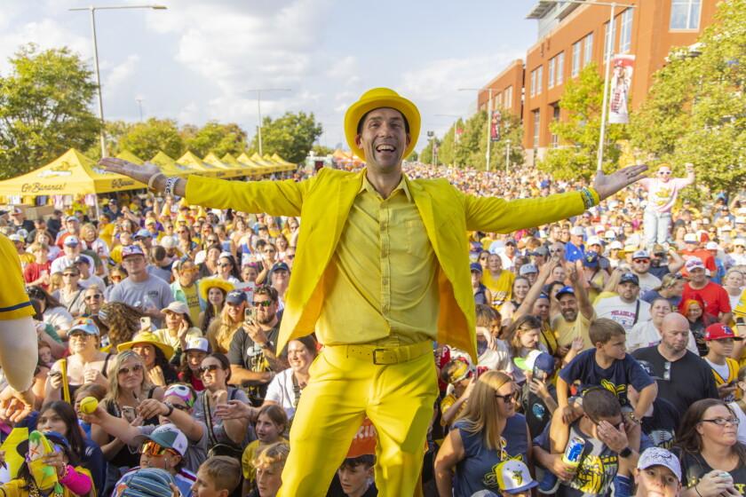 A performer in a bright yellow suit and hat stands with arms outstretched above a huge crowd of Savannah Bananas fans at a street festival